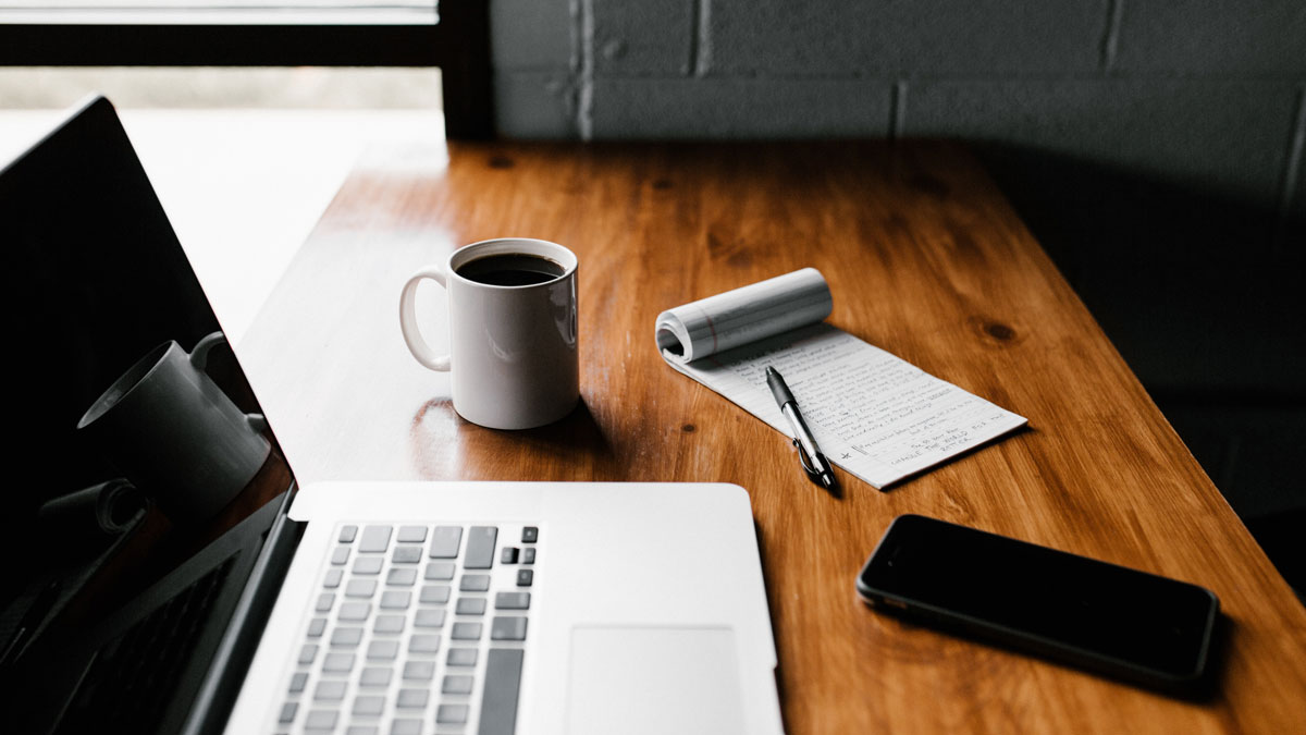 A laptop, phone, coffee mug and notepad sitting on a desk