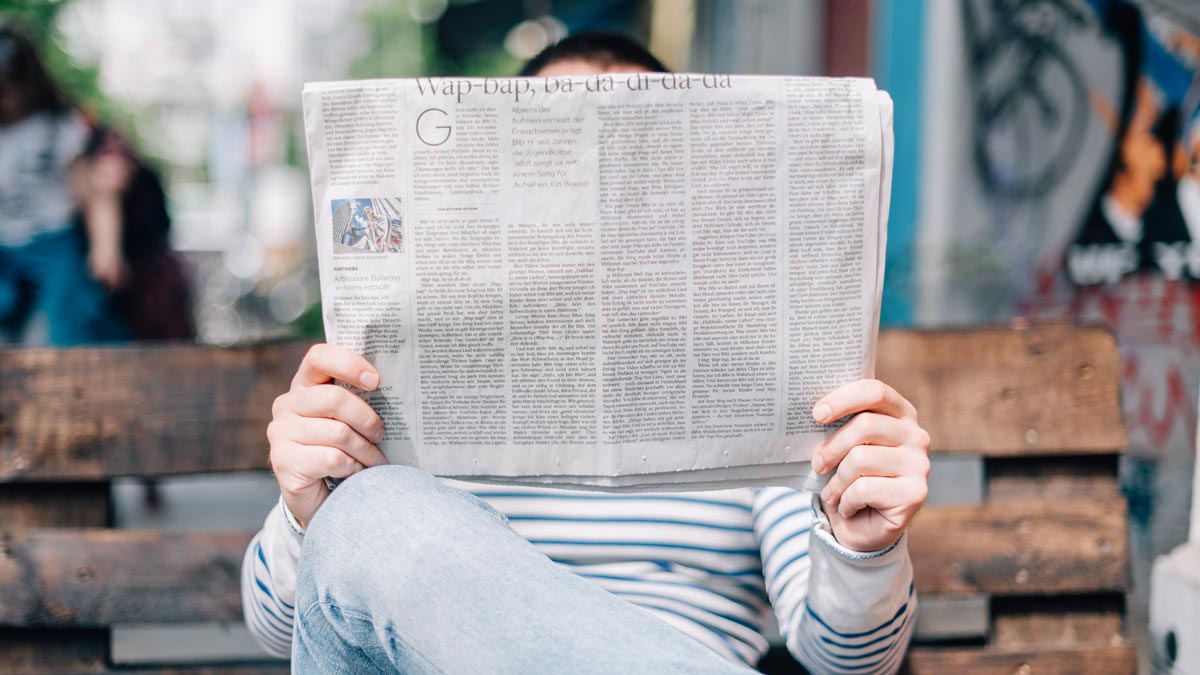 A person sits on a wooden bench, reading a newspaper